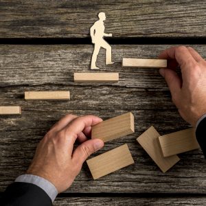 Silhouette cutouts of a man walking up steps, while the hand of other businessman building stairs for him in a conceptual image of personal and career promotion.